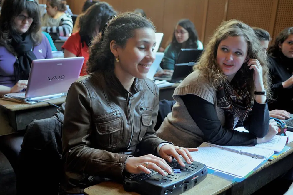 Une élève utilise une plage braille en classe, assise à un bureau aux côtés d’autres élèves travaillant sur ordinateur ou sur papier.