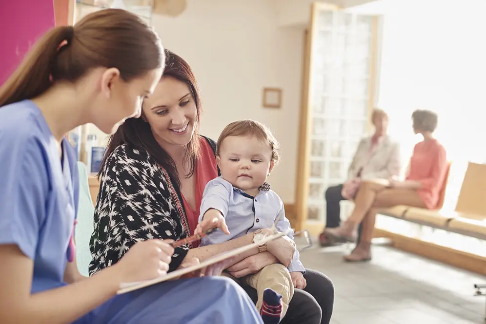 Femme portant un enfant main tendue vers le bloc note d'une femme soignante.