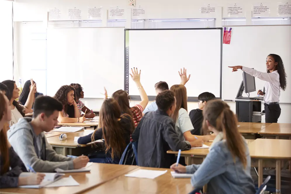 Élèves en cours levant la main pour répondre aux questions d'une enseignante dans une salle de classe.