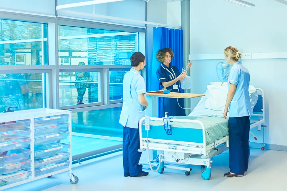 Trois jeunes femmes en blouse bleue autour d'un lit d'hôpital, l'une tenant une perfusion