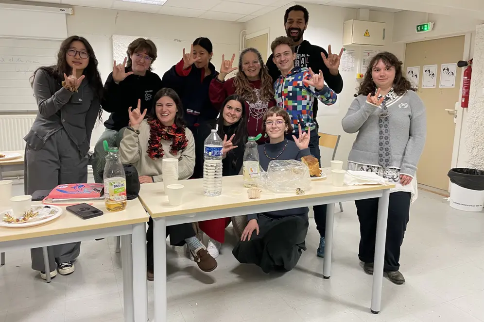 Un groupe de personnes pose dans une salle de cours autour de tables, en faisant le signe “je t’aime” en langue des signes.