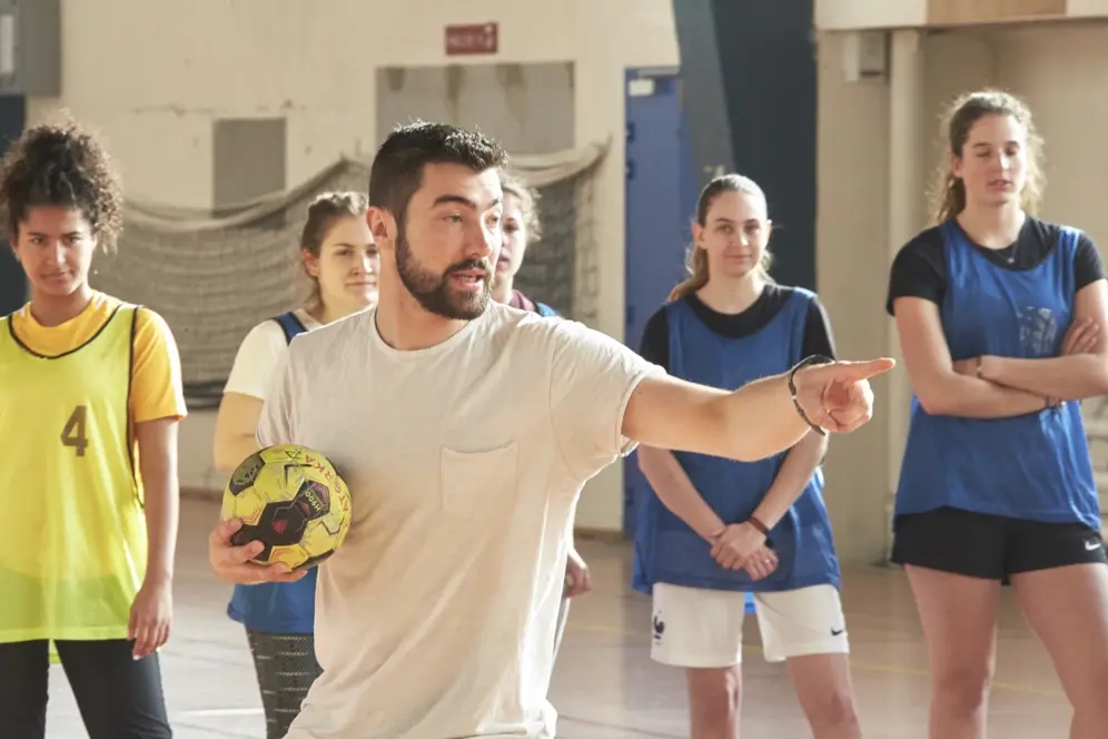 Homme tenant un ballon de handball et donnant des consignes par un geste de la main, entourée d’un groupe de joueurs dans un gymnase.