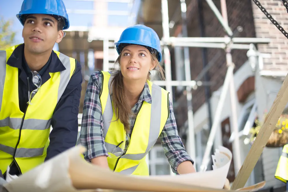 Une jeune femme et un jeune homme tous les deux casqués de bleu et vétus d'un gilet jaune consultent un plan sur un chantier