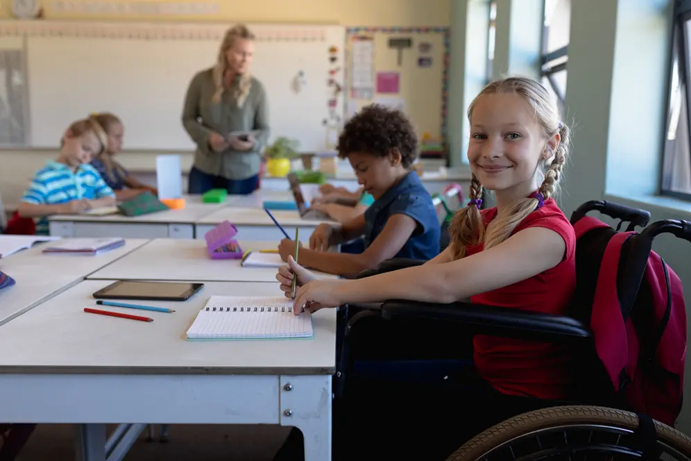 Photo d'une petite fille blonde et blanche en fauteuil roulant dans une classe d'école élémentaire.