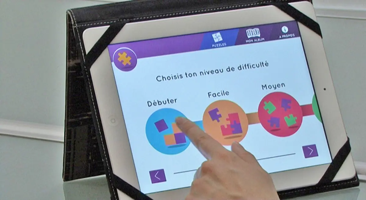 Vue sur un &eacute;cran de tablette et d'un index. On lit choisis ton niveau de difficult&eacute; puis 3 ronds color&eacute;s indiqu&eacute;s : d&eacute;buter, facile, moyen