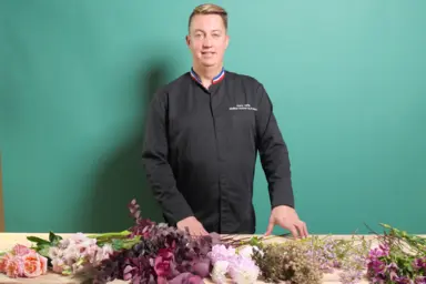 Professeur d'art floral avec ruban meilleur ouvrier de france debout derri&egrave;re un comptoir d&eacute;cor&eacute; de fleurs coup&eacute;es blanches, roses, mauves et vertes