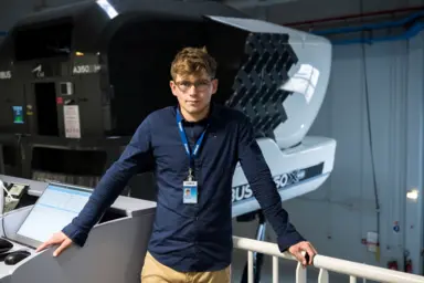 Portrait d'&Eacute;lio Lecellier, jeune gar&ccedil;on blanc et blond. Il se tient adoss&eacute; &agrave; une barri&egrave;re, un ordinateur &agrave; sa droite, du mat&eacute;riel d'aviation d'Airbus derri&egrave;re lui.