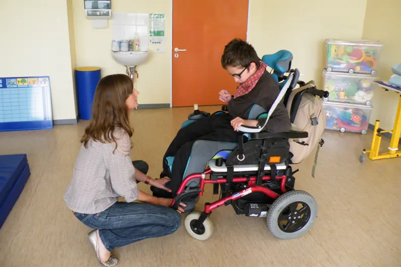 Gwendoline, la psychomotricienne, est accroupie face &agrave; Alexandre, son patient, assis dans son fauteuil roulant. Ils discutent.