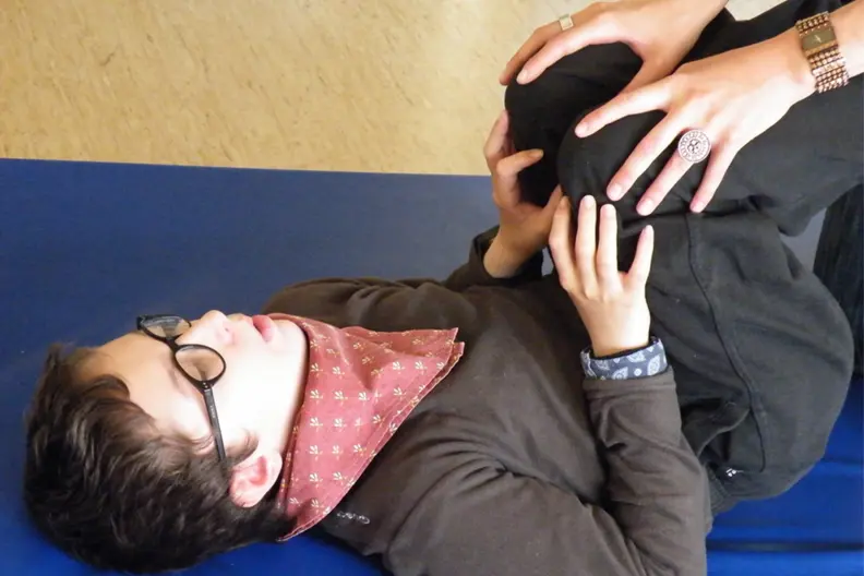 Un gar&ccedil;on blanc et brun est allong&eacute; sur un tapis bleu. Ses genous sont repli&eacute;s contre son torse. Une adulte l'aide &agrave; les maintenir dans cette position.