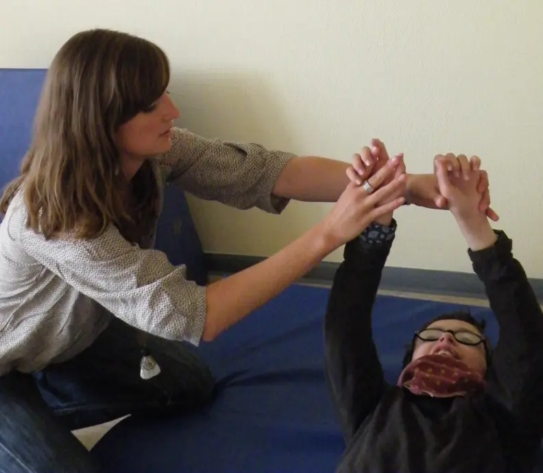 La psychomotricienne aide Alexandre &agrave; faire des mouvements de bras. Il est allong&eacute; sur un tapis bleu et elle est &agrave; genous &agrave; c&ocirc;t&eacute; de lui.