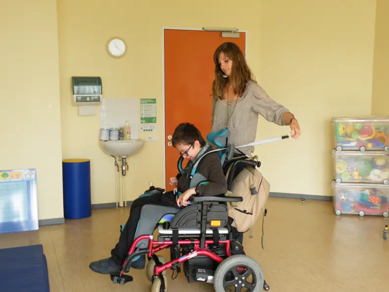 Gwendoline, la psychomotricienne, fait travailler Alexandre, assis dans son fauteuil roulant, pour sa s&eacute;ance de yoga. Ils sont dans une salle m&eacute;dicale aux murs jaunes.