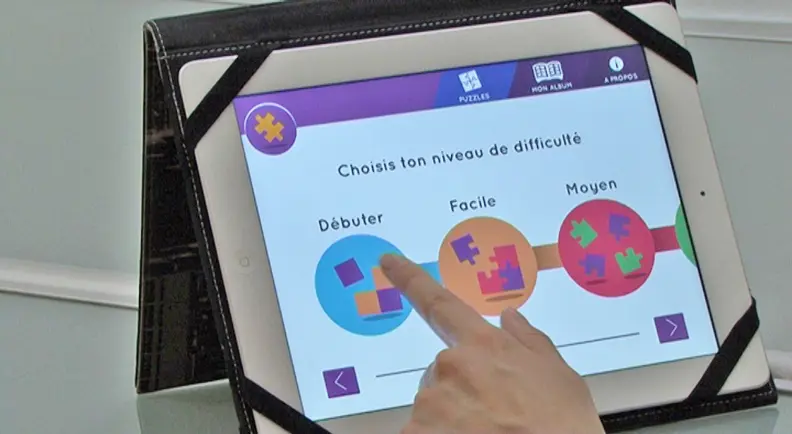Vue sur un &eacute;cran de tablette et d'un index. On lit choisis ton niveau de difficult&eacute; puis 3 ronds color&eacute;s indiqu&eacute;s : d&eacute;buter, facile, moyen