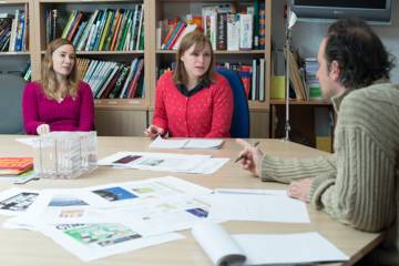 2 femmes et un homme discutent autour d'une table de travail remplie de papier