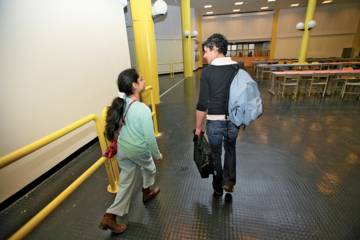 Deux personnes marchent dans un bâtiment scolaire, l’une portant un sac à dos et l’autre un sac en bandoulière, dans un espace avec tables, chaises et structures jaunes.