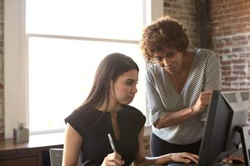 Apprentie devant un ordinateur à son poste de travail, accompagnée par son maître d'apprentissage dans une entreprise.