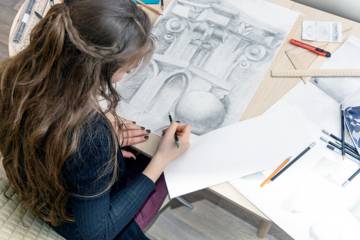 Une jeune femme dessine sur une table couverte d’outils et de croquis, concentrée sur un travail de dessin technique.