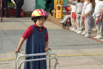 Une enfant avance avec un déambulateur sur une cour d’école, portant un casque coloré, tandis que d’autres enfants attendent en ligne près des structures de jeu.